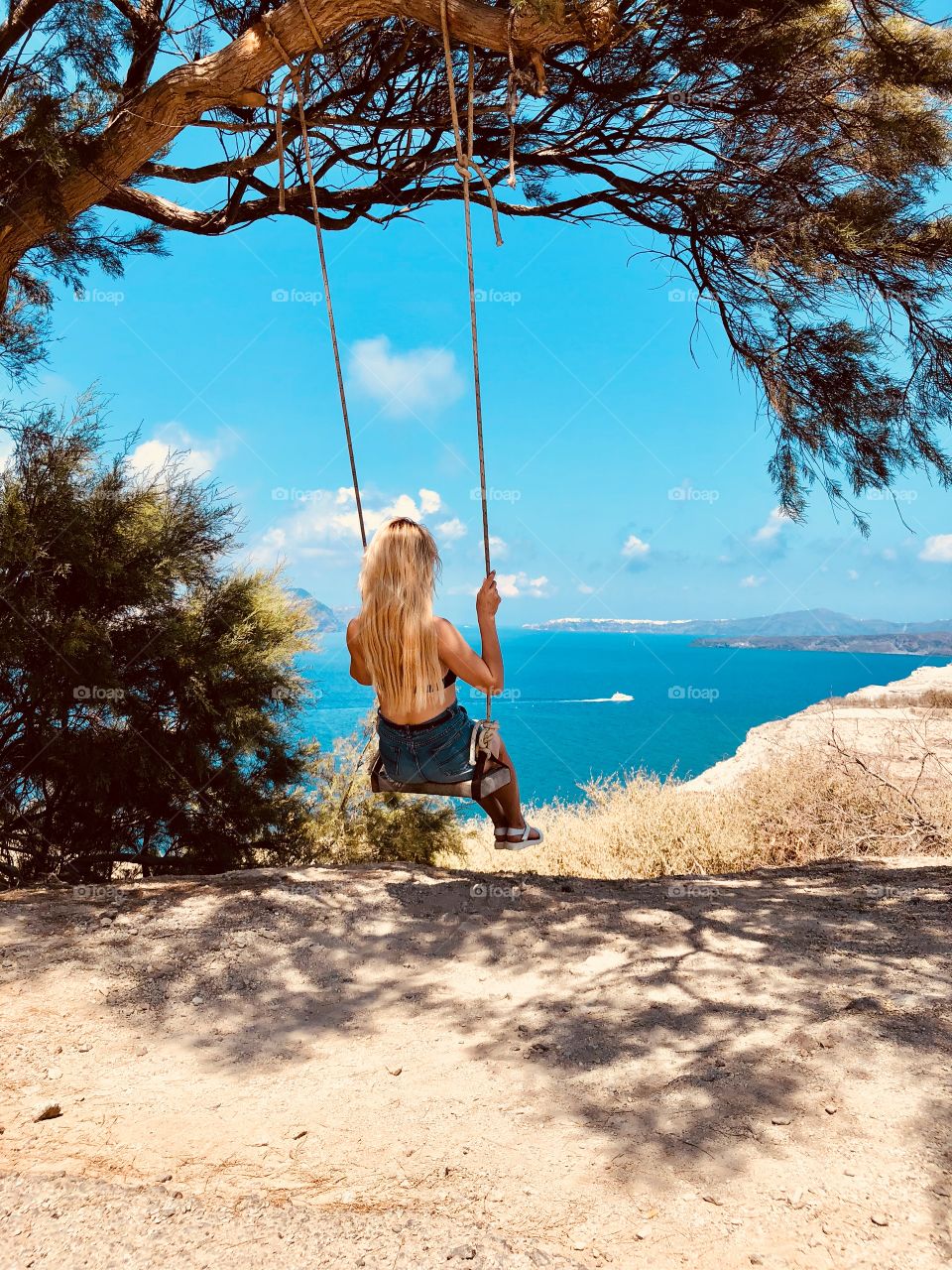 A young blonde girl swinging on a swing, around the beautiful views of the sea and the cliffs of Santorini Island.