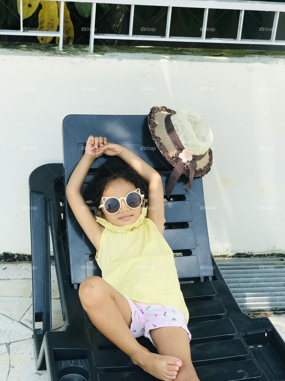 The baby girl in summer outfits and cooling glasses to her eyes lean over the pool lounge chair and relaxing 