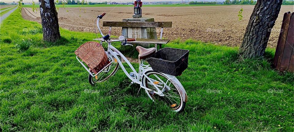 This is an idyllic rest stop with a bench and a crucifix under a small symbolic triangle roof above it in rural “Niederbayern” or “Lower Bavaria”, Germany. Our photographer has leaned her new bicycle against it. 2024. Hypnotic Productions