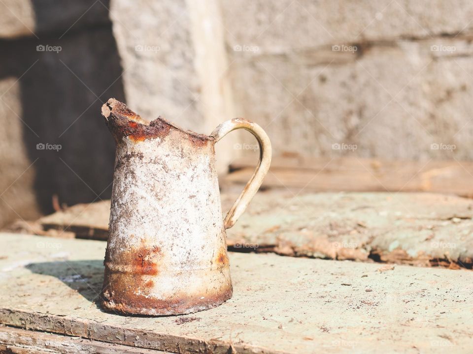 One old rusty and rotten enameled jug with a handle stands on planks on a concrete wall in the back yard of the house, close-up side view. The concept of retro objects, vintage crockery, metal.