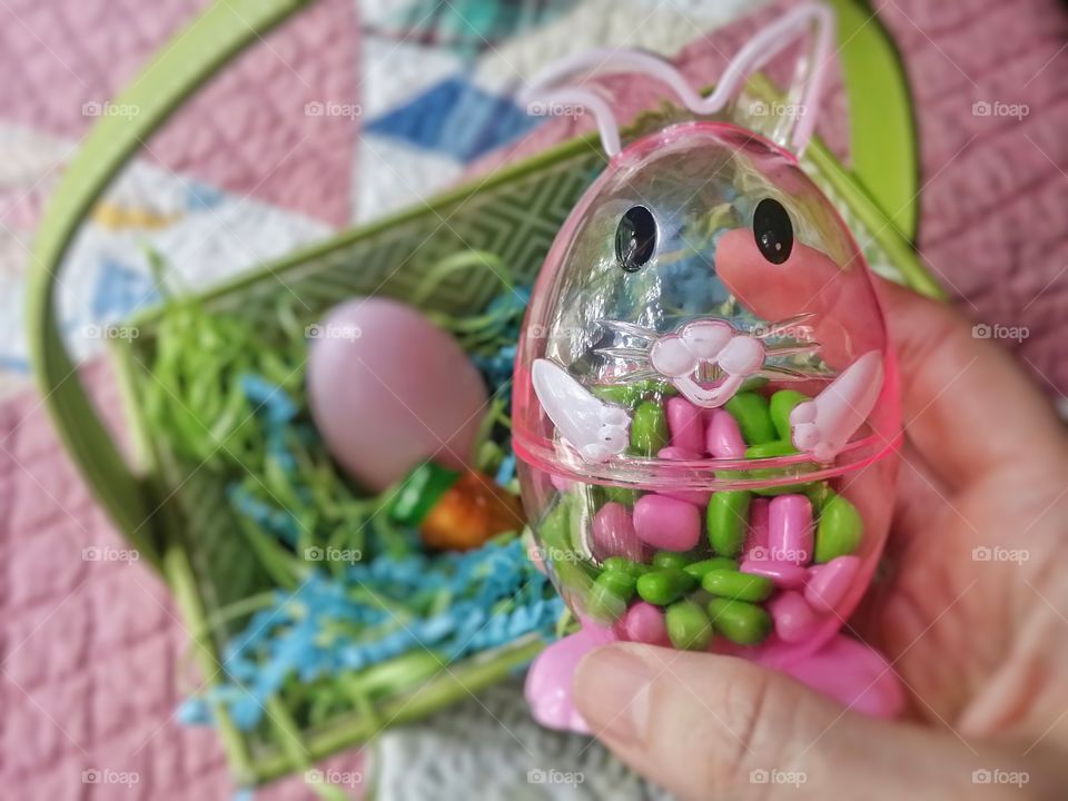 A clear pink Easter bunny egg filled with pink and green candy being held by a hand with an Easter basket with a plastic egg and chocolate carrot sitting on a quilt in the background