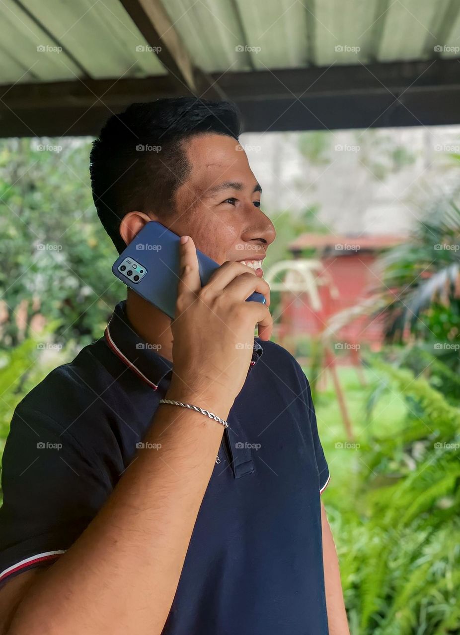 Young dark-skinned man smiling while talking on a light blue cell phone in a park with a garden