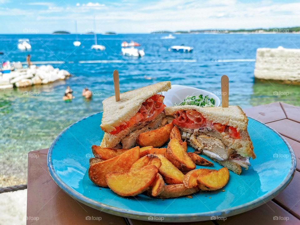 Close-up photo of club sandwich with potatoes on blue plate in a beach bar