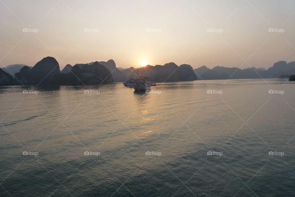 On the water there are 2 yachts , the water is slightly rippling. Surrounding there are many undulating mountains, the sun is falling,shines into the water,creating beautiful light at sunset