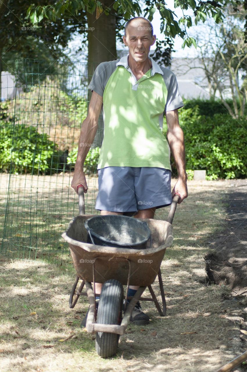 middle-aged gardener in dirty work clothes pushing a bucket cart, rusty metal cart for garden work, sunny spring day, work mood, start work in spring in the garden