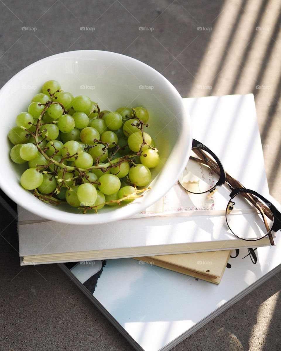 A stack of books, notebook, glasses, a plate of green grapes on a concrete table, nobody 