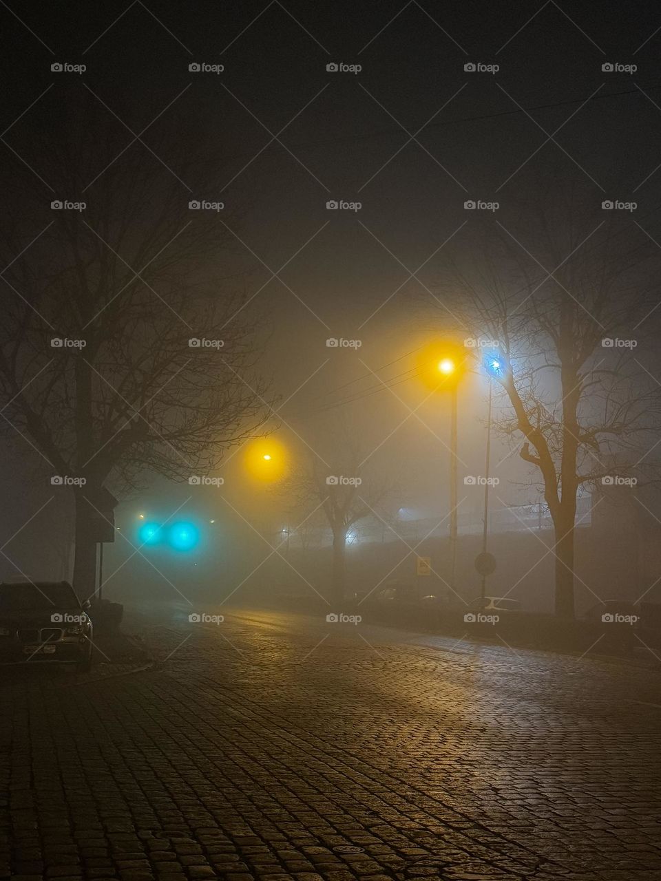 Foggy evening, empty street with no people, lonely car on the side of the road, warm lantern lights and green traffic lights 