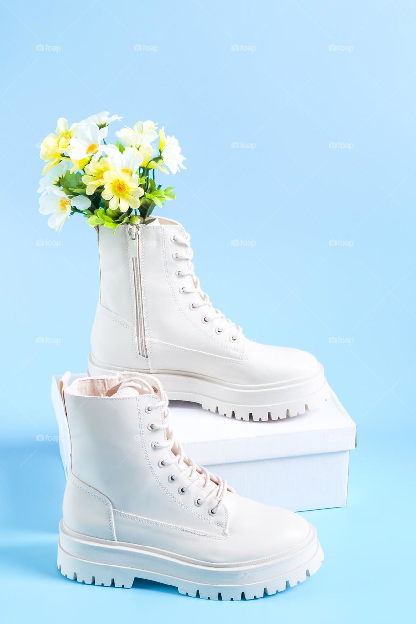 One pair of beautiful white Martens brand shoes on a rough tractor sole with lacing and an artificial bouquet of spring flowers stand on a white cardboard box in the center on a light blue background,side view. The concept of fashionable women's shoe