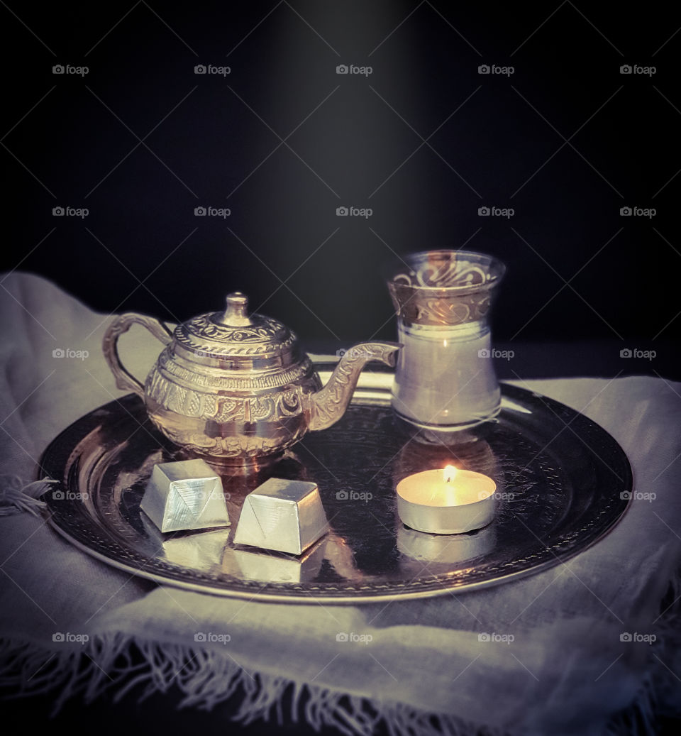 Dark oriental still life. Silver ornamental tray with a small silver teapot and a glass of tea with milk, sweets and a burning candle on gray beautiful textile. Tasty snack. Food photo, square orientation