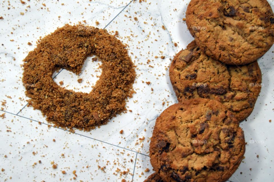 A simple heart hollowed out of the centre of a sprinkling of cookie crumbs sits beside a pile of chocolate chip cookies.
