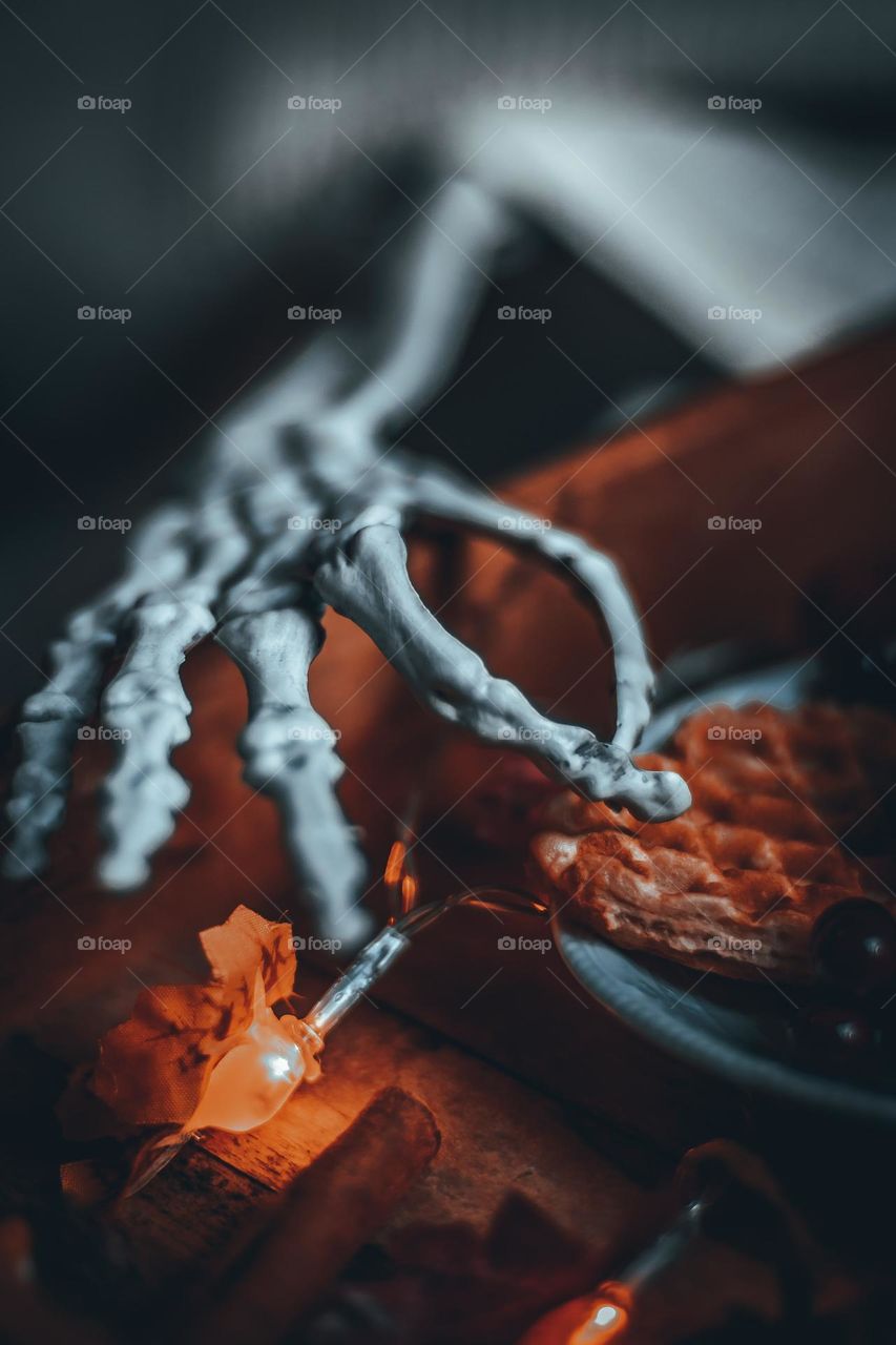 One arm of the skeleton sticking out from under the blanket on the bed reaches for a crayfish in a wooden tray standing on the bedside table, side view, close-up. Halloween breakfast concept, holiday food.