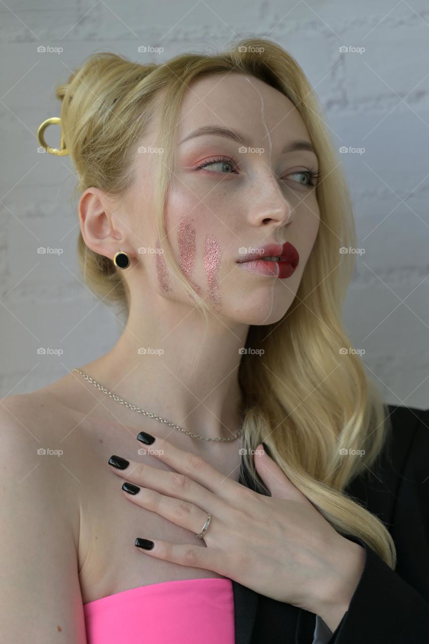 Close-up portrait of a blonde girl with different make-up on half of her face and a thoughtful look with her hand on the chest on white background 