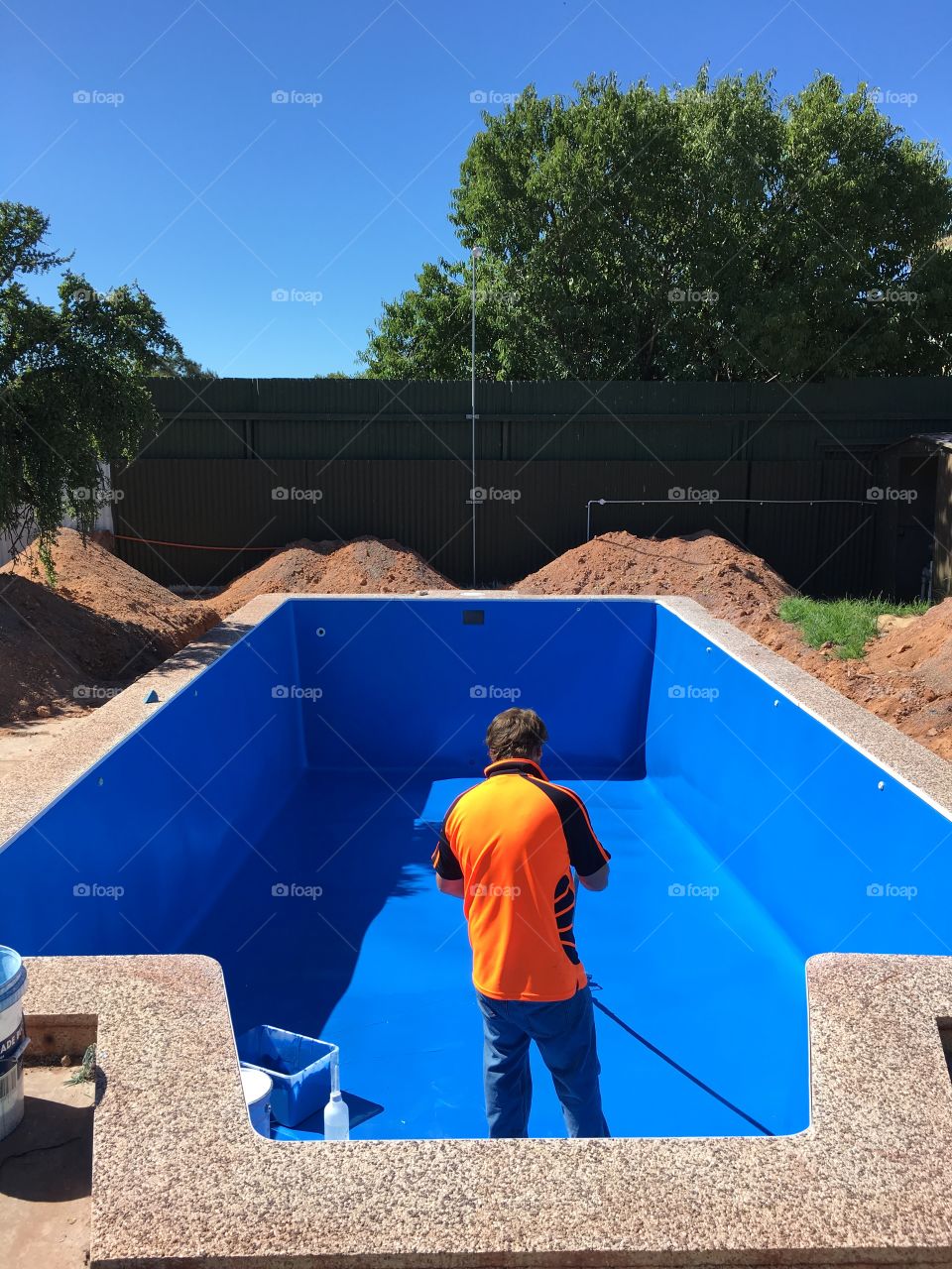 Man in orange shirt working on empty outdoor swimming pool painting the fibreglass liner