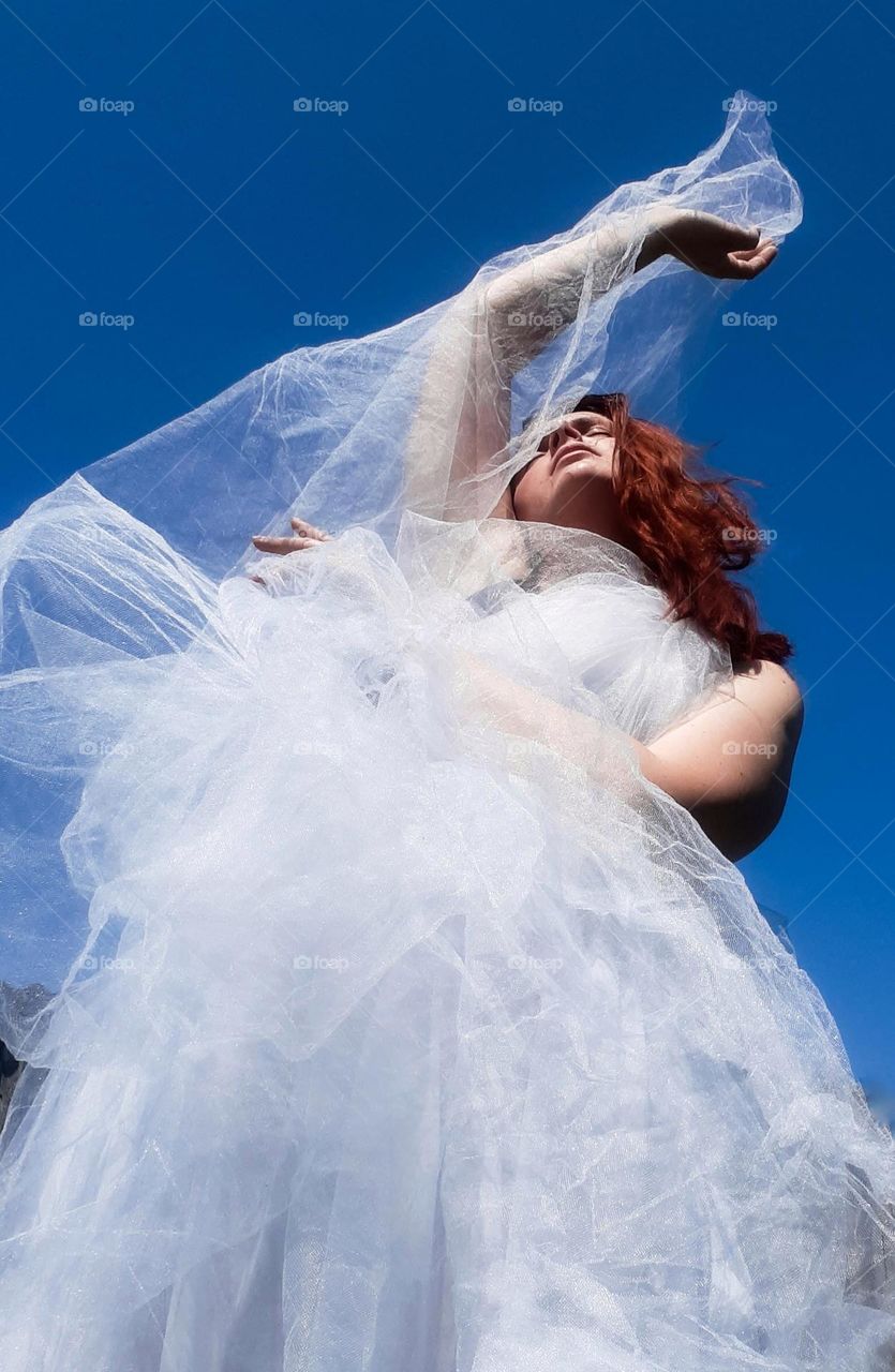 original contrast portrait of a red-haired girl in a white satin dress against a clear blue sky