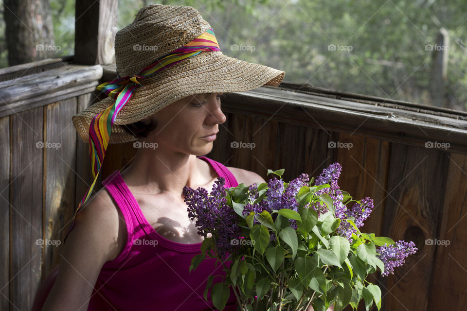 A woman with summer hat on a balcony of country house holds lilac flowers