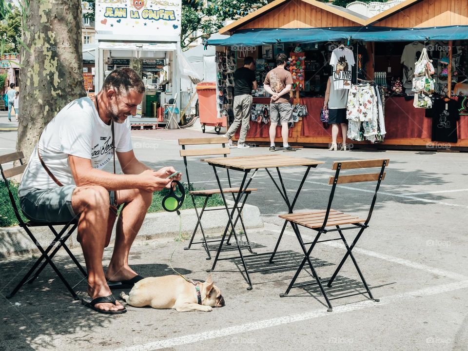 One handsome young and happy man sits at a table of a street cafe laughing looking at the screen of a mobile phone, holding his dog on a leash, close-up side view.