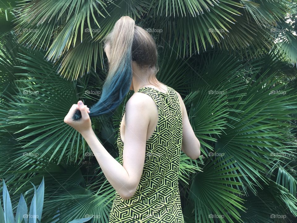 A girl standing back to the camera palyong with her hair. In the background there is a big plant