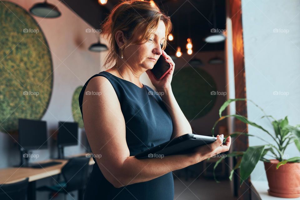 Businesswoman having business call working on tablet in office. Mature busy woman using touch pad computer standing by window in modern interior. Female manager focused on work holding digital device. Using technology