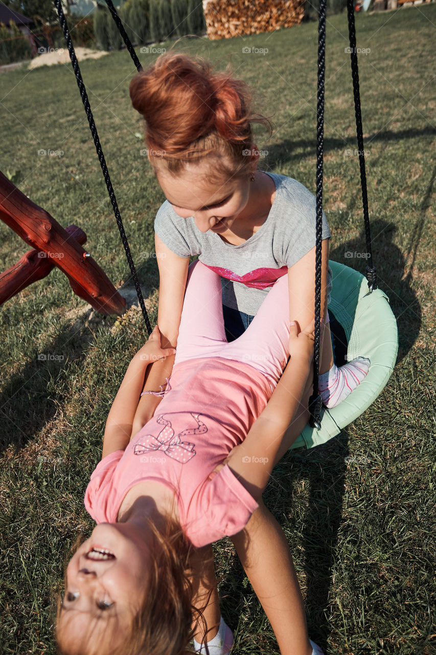 Teenage girl playing with her younger sister in a home playground in a backyard. Happy smiling sisters having fun on a swing together on summer day. Real people, authentic situations