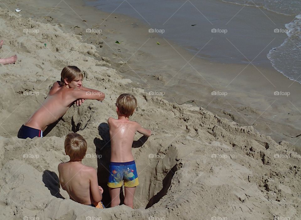 A group of boys enjoying a day at the beach in holes they dug in the sand in Folkestone on the coast of Southeast England on a sunny summer day. 