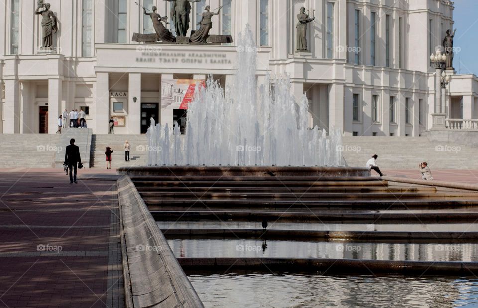 Fountain in the city, near the theater, people rest near the water.