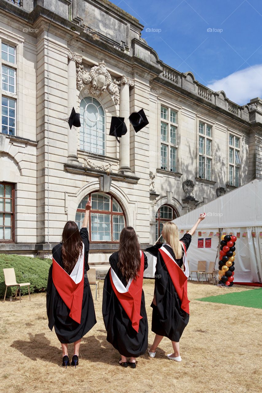 Back side view of girls wearing academic dresses and throwing academic caps in yard of university 