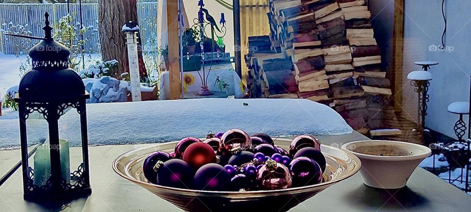 In a house in „Bavaria“, Germany a  bowl filled with Christmas decorations, shiny balls of various sizes, a lantern, candelabra and outside firewood stacked against a wall all the way to the ceiling can be seen. 2022. Hypnotic Productions