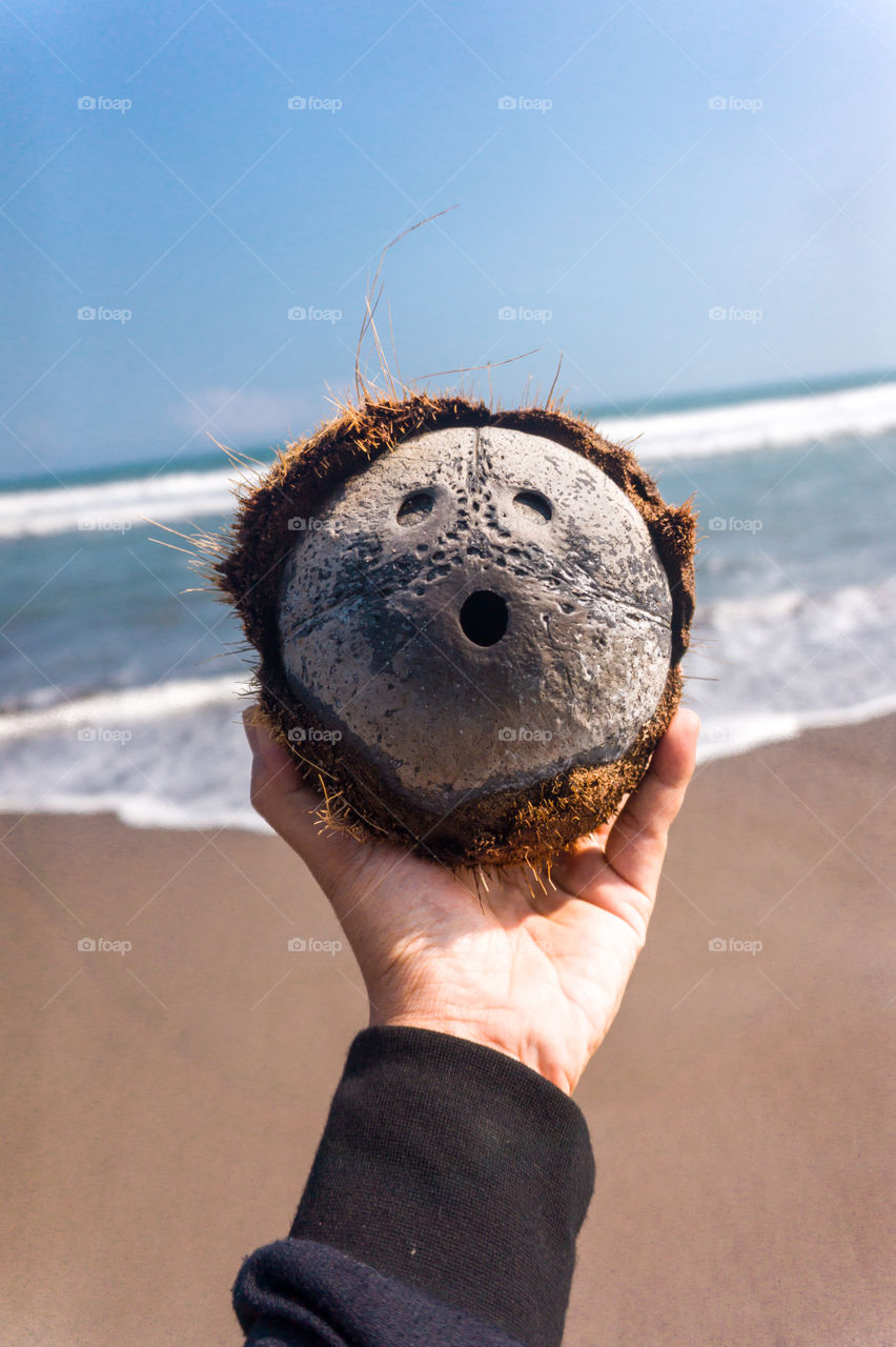 Close-up hand holding coconut shell that resembles a human face with beach background