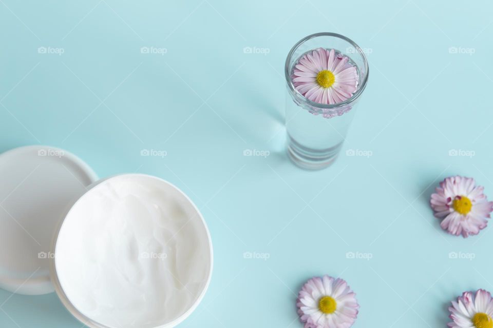 daisy in a glass of water on a blue background next to a jar of white cream