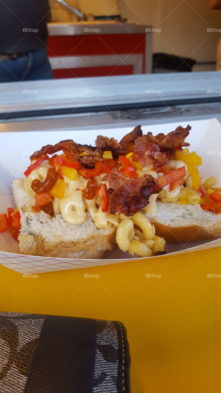 Hotdog covered in macaroni, sliced bacon, red pepper and yellow pepper.