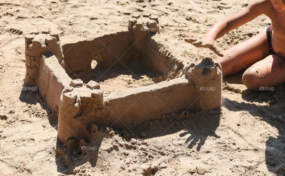 One little Caucasian unrecognizable girl builds a sand castle on the shore near the sea on a summer sunny day, close-up side view.