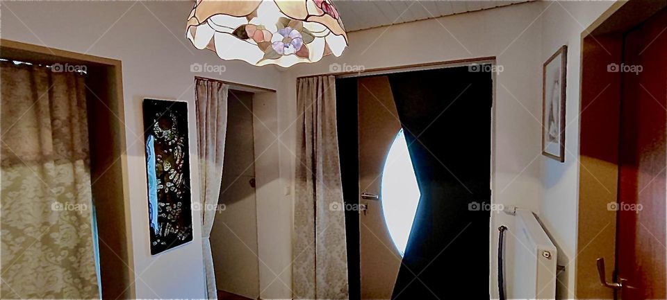This is the view of the „Art Deco“ style front door from the inside of the beautiful one story house in „Bavaria“, Germany. Thermo curtains provide added insulation. A beautiful „Tiffany“ style lamp hangs from the ceiling. 2023. Hypnotic Productions