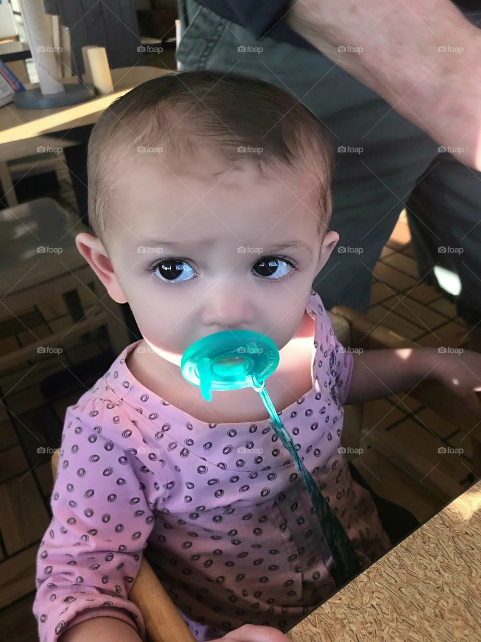 Photo of concerned baby girl with giant brown eyes and green pacifier sitting at the table waiting to be fed