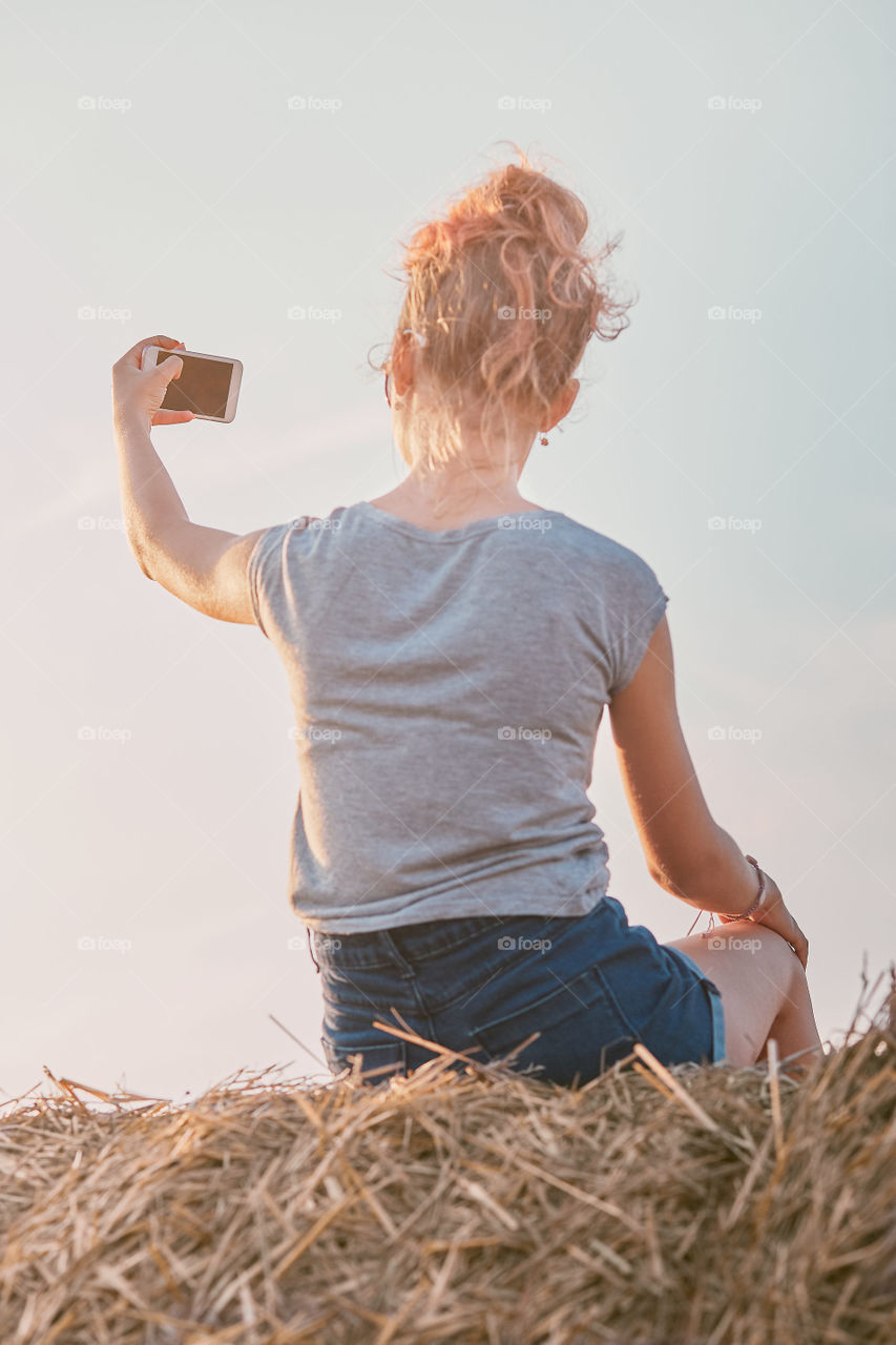 Teenage girl taking a selfie of herself, sitting on a hay bale enjoying summer vacation in the countryside. Candid people, real moments, authentic situations