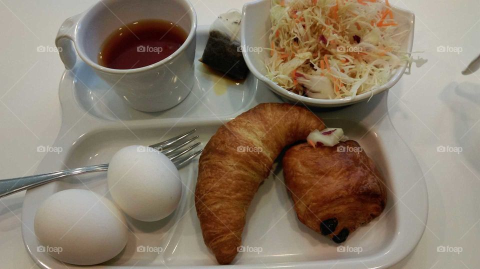 one bread,one croissant, 2 boiled eggs,one bowl of salad and a cup of tea, served on the white tray,special breakfast complete with nutrition.