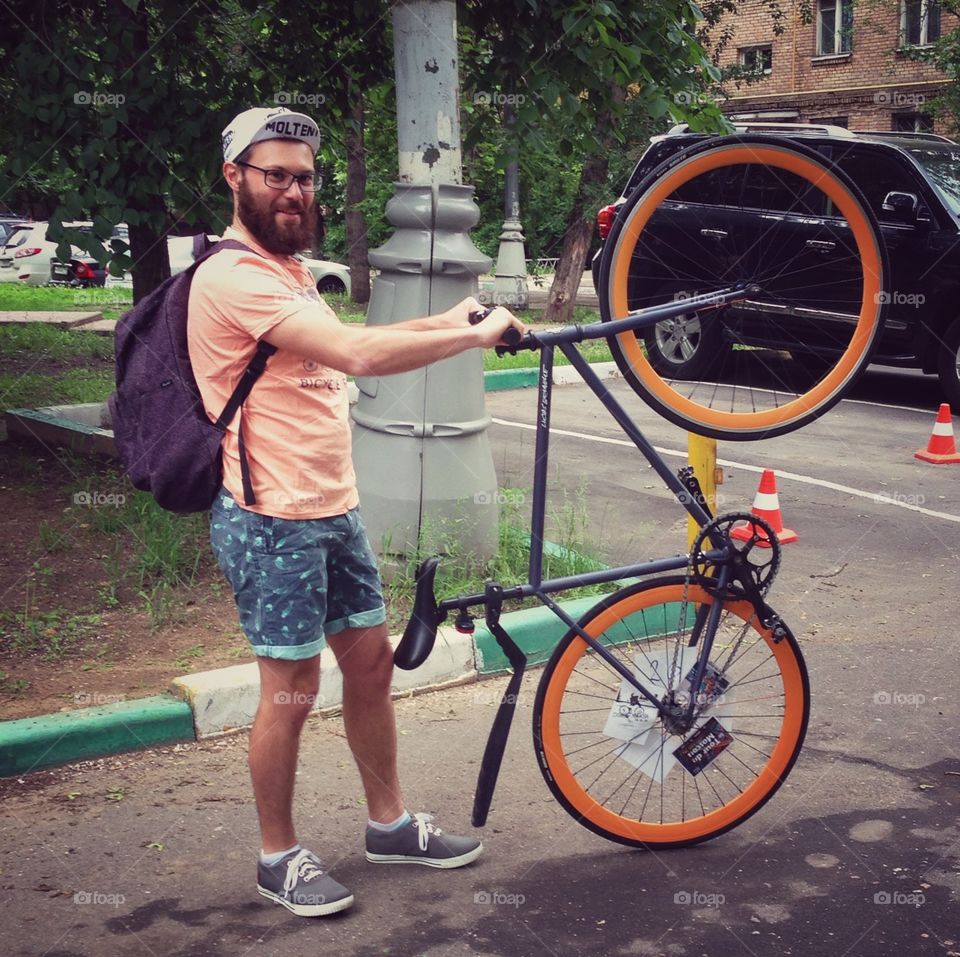 Man with beard and glasses in pink T-short, cycling cap, backpack and shorts with grey brakeless fixie bike with orange wheels