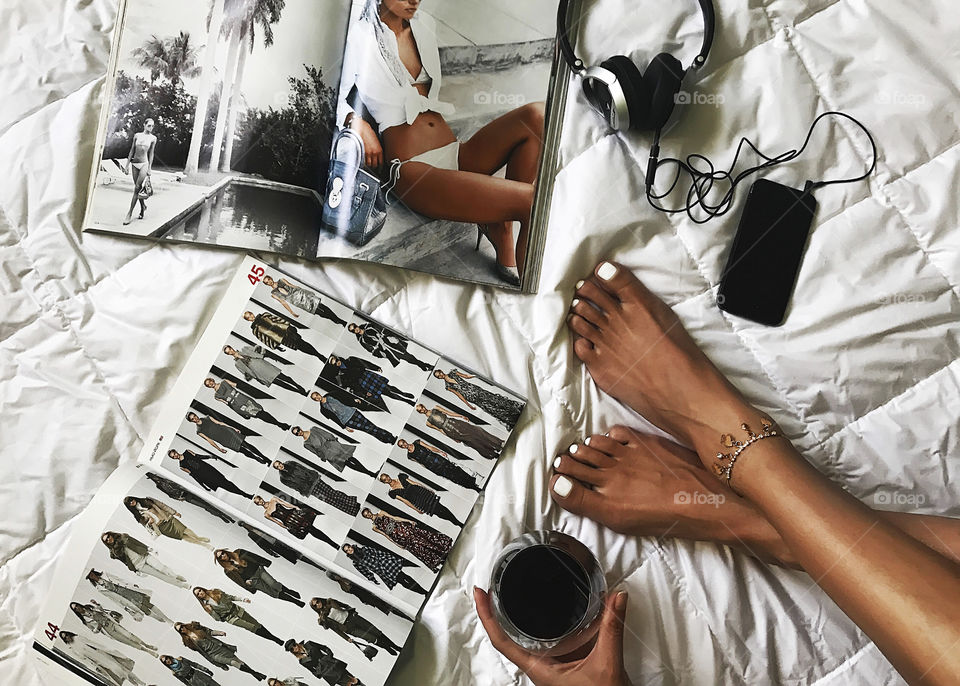 Overhead view of a home relaxing at cozy bed - reading fashionable magazines, music listening, drinking red wine 