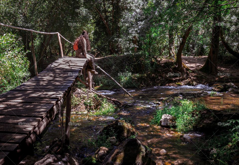 A hiker takes a break sitting on a bridge over a small stream in woodland