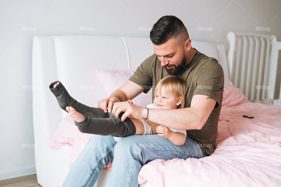 Young happy family with father dresses daughter baby girl on bed in bright room at cozy home