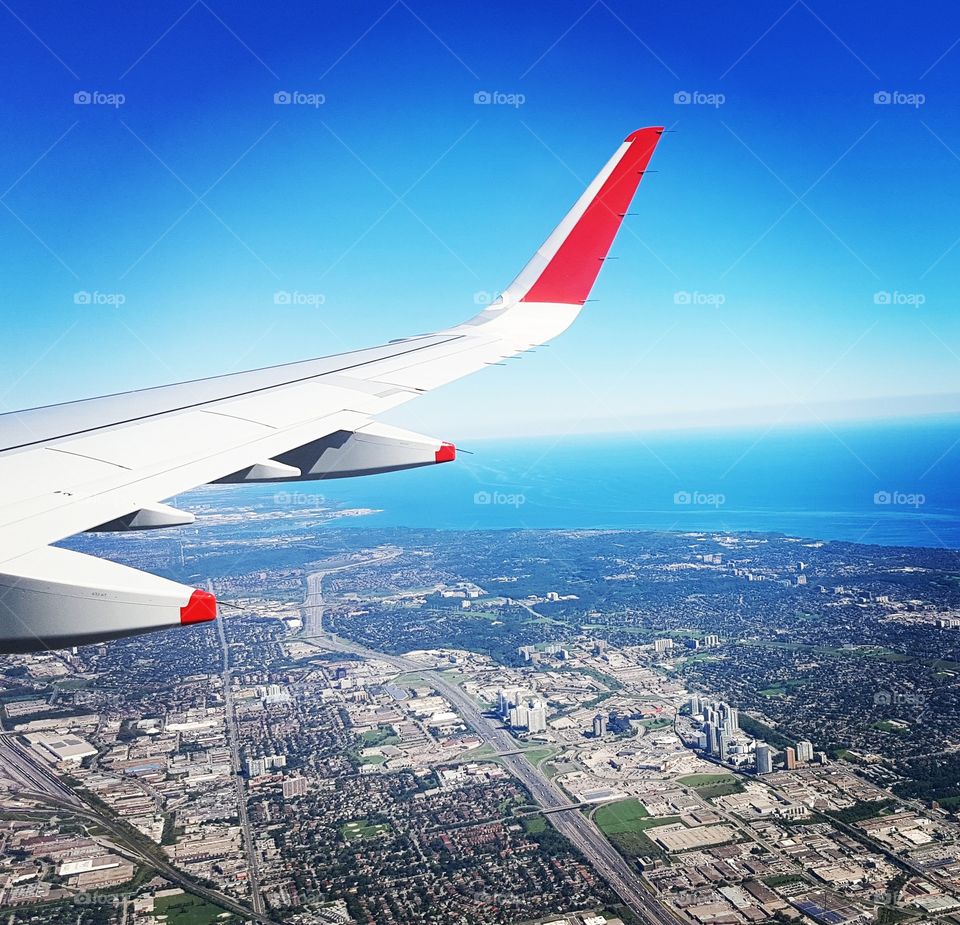 red and white aircraft wing and aerial city view with ocean below flying through cloudless blue skies