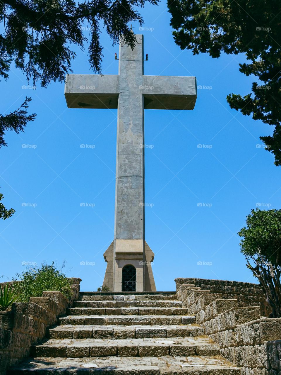 A beautiful view of a large stone cross with an old stone staircase to it along a cypress alley on Mount Filerimos in Greece, close-up from below.