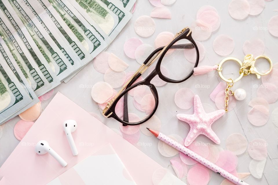 Flatlay items: cash money one hundred dollars bill, reading glasses, wireless headphones, notebook, pencil, star pink color, confetti lying on gray concrete background 