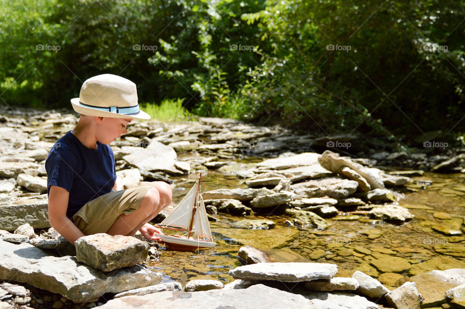 Young boy playing with a toy sailboat in the water of a creek during the summer