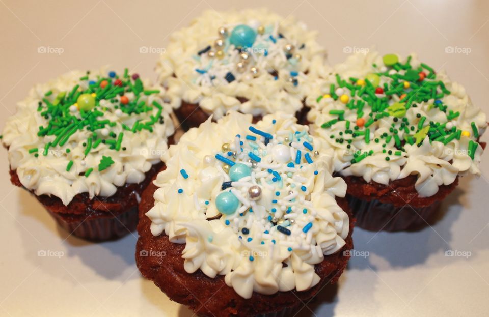 Christmas baking time! A closeup of 4 red velvet cupcakes with vanilla icing and blue, white, silver, gold, green, red, yellow, sprinkles, dragees and little Christmas trees. Yummy!