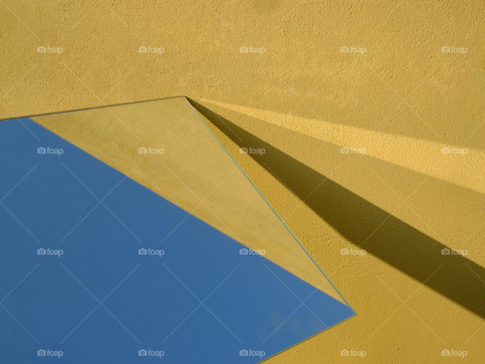 An abstract view of a mirror reflection of the blue sky against a yellow painted wall