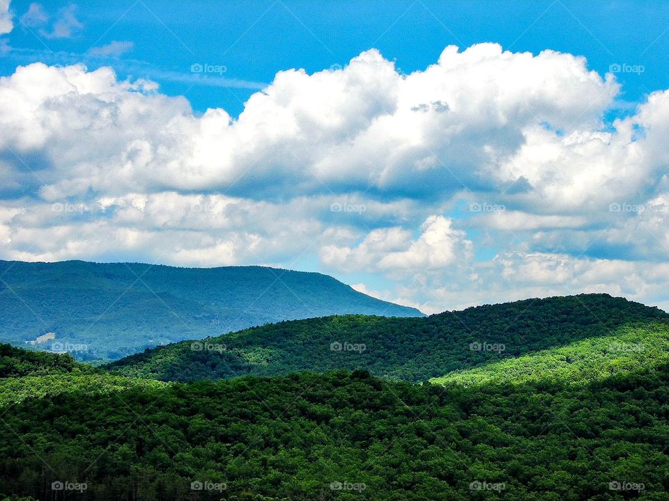 Scenic landscape with rolling hills, green trees, white clouds, and blue sky.