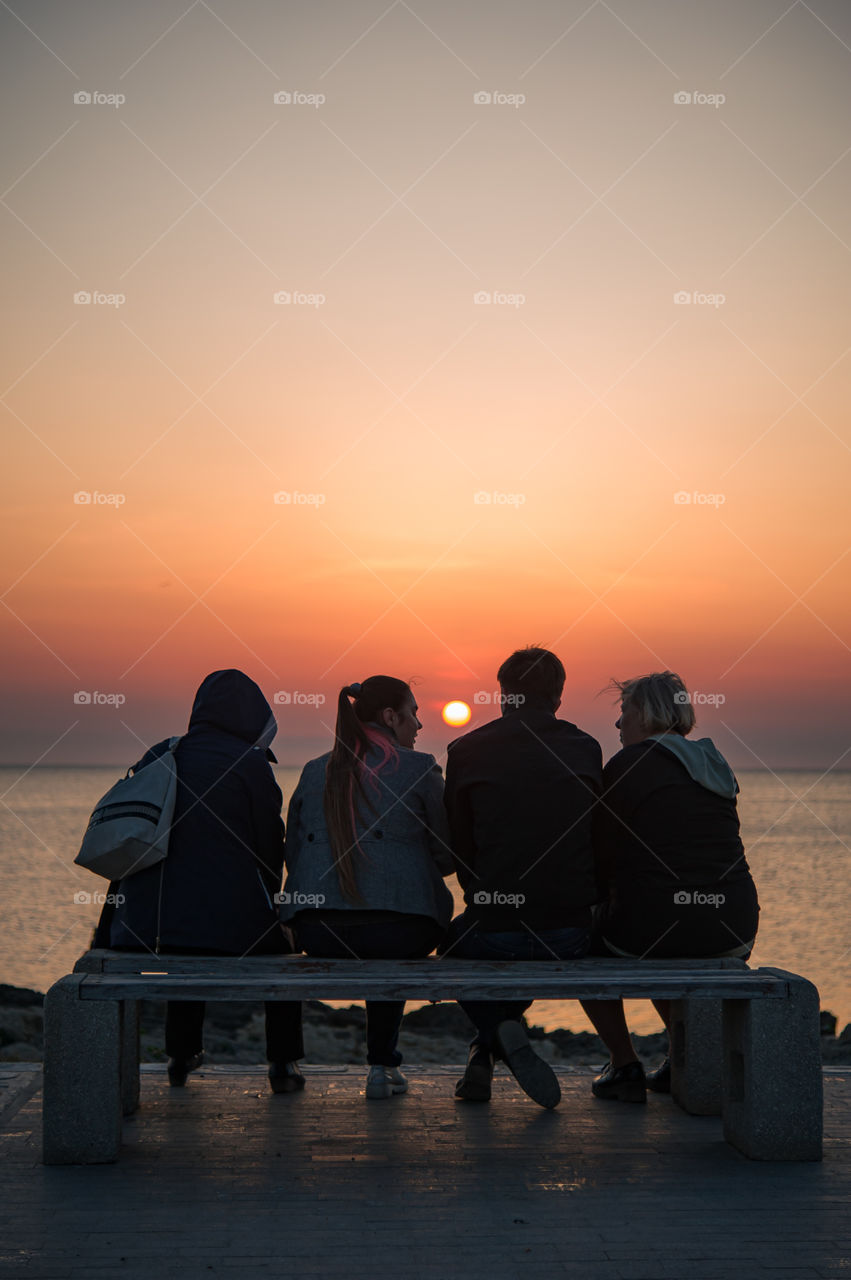 A family of four sit on a bench on the Black Sea, communicate and enjoy the beautiful sunset.