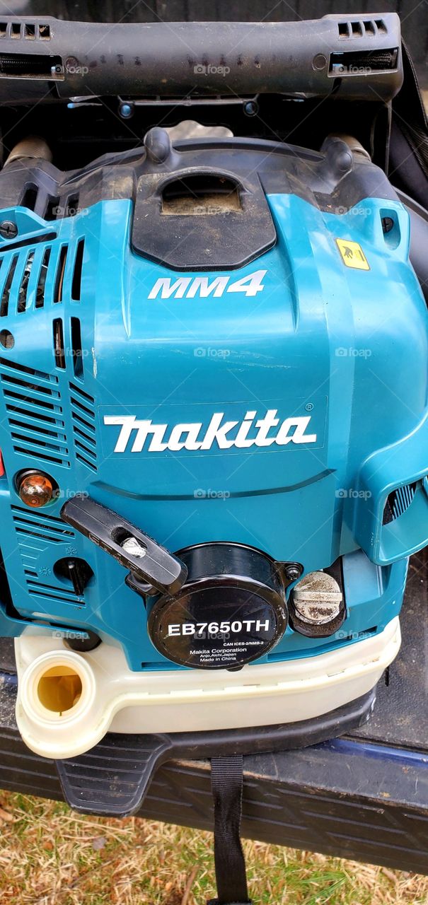 Blue Makita backpack motor for leaf blower. There is a pull start gasoline motor with gas tank cover off for refilling.