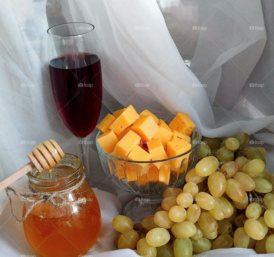 Yellow color.  On a white background, a composition of a glass with red wine, a vase with yellow cubes of Cheddar cheese, a bunch of yellow-green grapes and a jar of dark yellow honey, on which lies a wooden spoon, from which honey flows