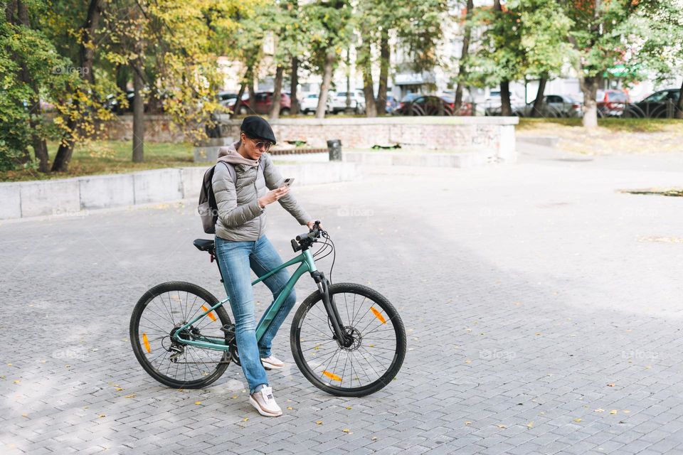 Portrait of fashionable young pretty woman in cap and sunglasses using mobile on bicycle on sunny autumn day in city park